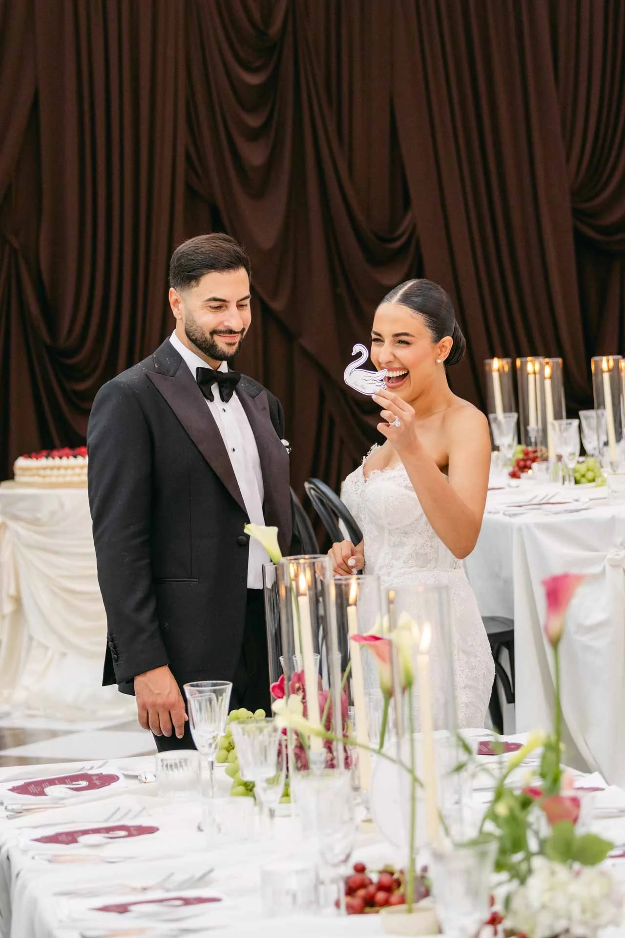 Bride and groom at their wedding reception, standing at a decorated table with flowers and candles. The bride is laughing and holding a swan-shaped cake topper. The groom is wearing a black tuxedo.