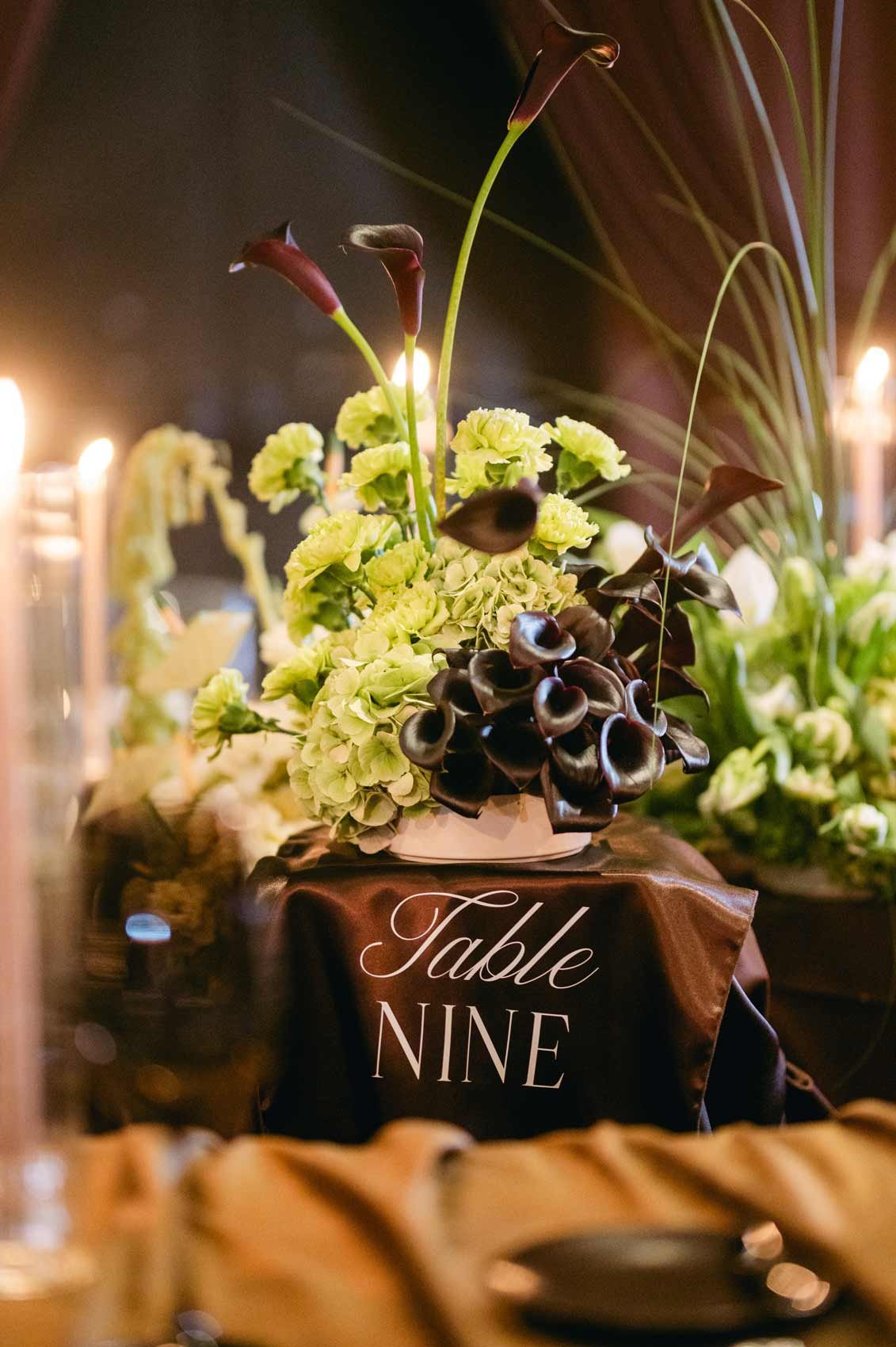 A floral centerpiece with dark calla lilies and light green hydrangeas on a table labeled 'Table Nine', inside a dimly lit setting.