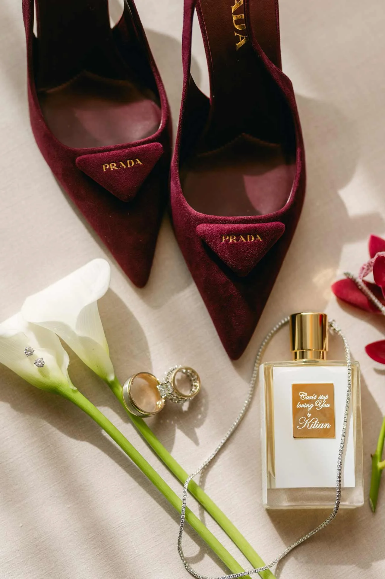 Burgundy Prada high heels, white calla lilies with diamond earrings, a gold ring, a personalized perfume bottle, and a silver necklace on a light-colored surface.