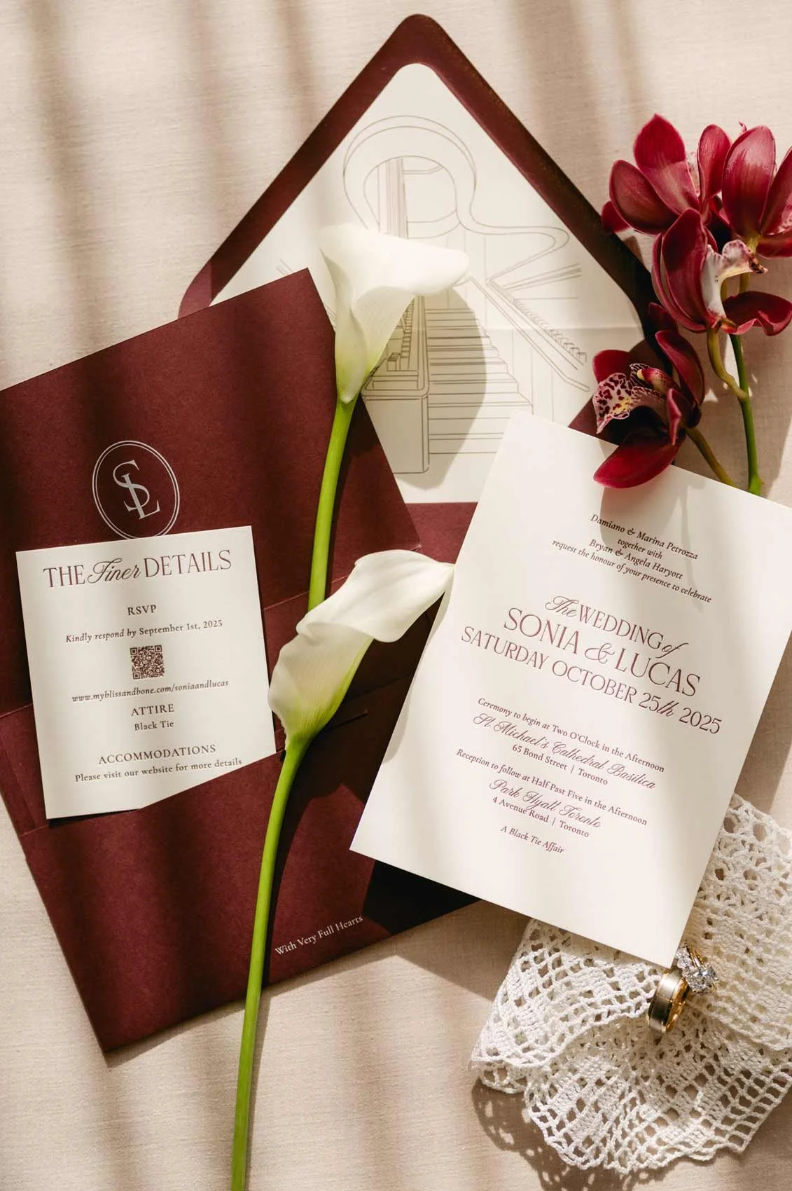 Wedding invitation card for Sonia and Lucas with details about the ceremony and reception, surrounded by a white calla lily flower, a red orchid, and a lace fabric with wedding rings placed on it.