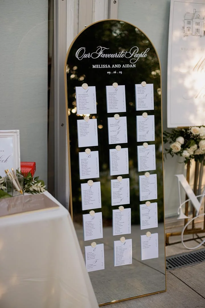 Wedding seating chart with guest names organized by tables, titled "Our Favourite People" with the names Melissa and Aidan and the date 09-16-23, displayed on a standing mirror board.