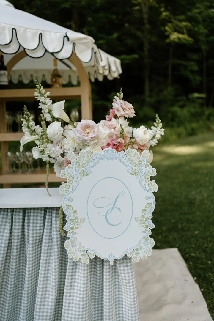 A decorative cake table with a white and gray checked tablecloth, a floral arrangement of pink and white flowers, and an ornate sign with the letter 'C' in script, outdoors with trees in the background.