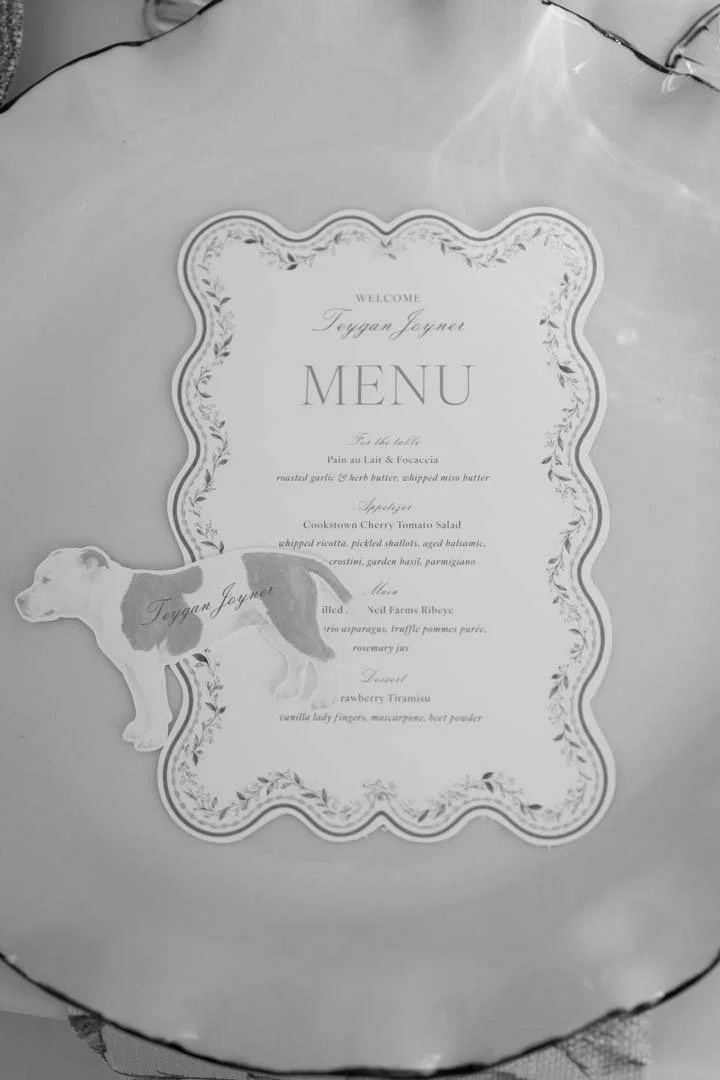 Wedding menu card with decorative border, floral design, and a dog-shaped name tag, on a table surface.
