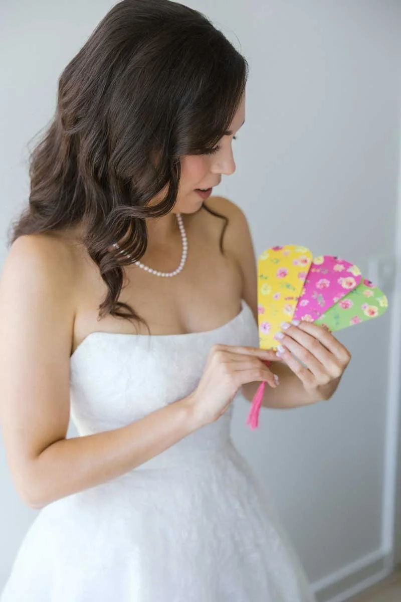 A woman with wavy brown hair wearing a white strapless dress and pearl necklace is holding four colorful floral-patterned fans.