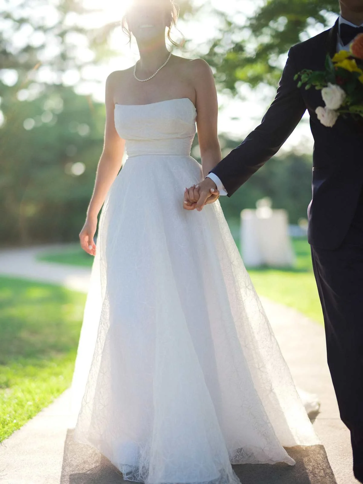 A bride and groom holding hands outdoors during their wedding, with sunlight shining through trees in the background. The bride is wearing a strapless white dress and pearl necklace, and the groom is in a black tuxedo with a bouquet of flowers.
