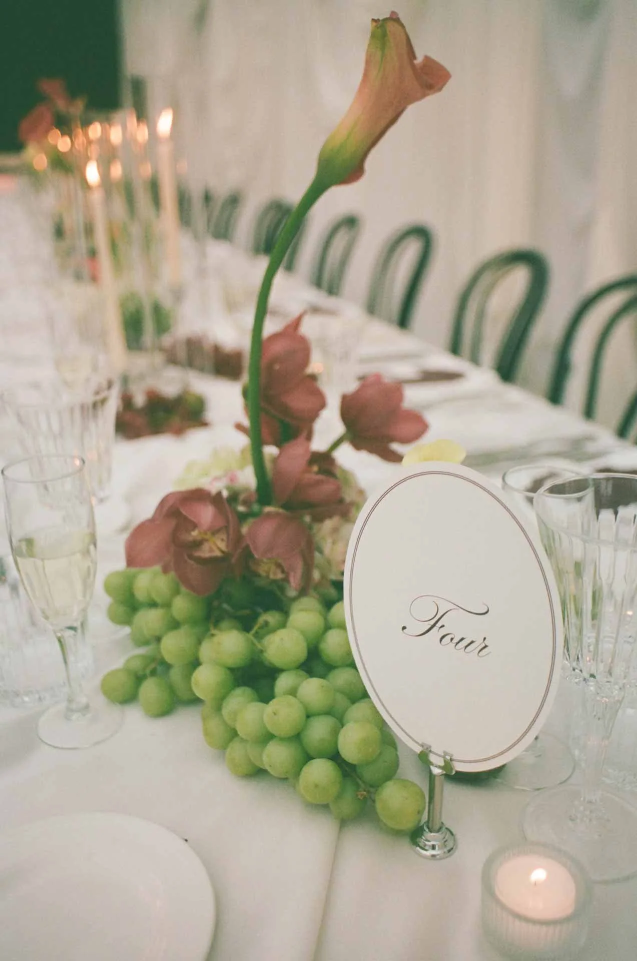 Elegant table centerpiece featuring a grapevine with green grapes and pink flowers, accompanied by a table card with the word 'Four' and lit candles, set for a formal event or dinner.
