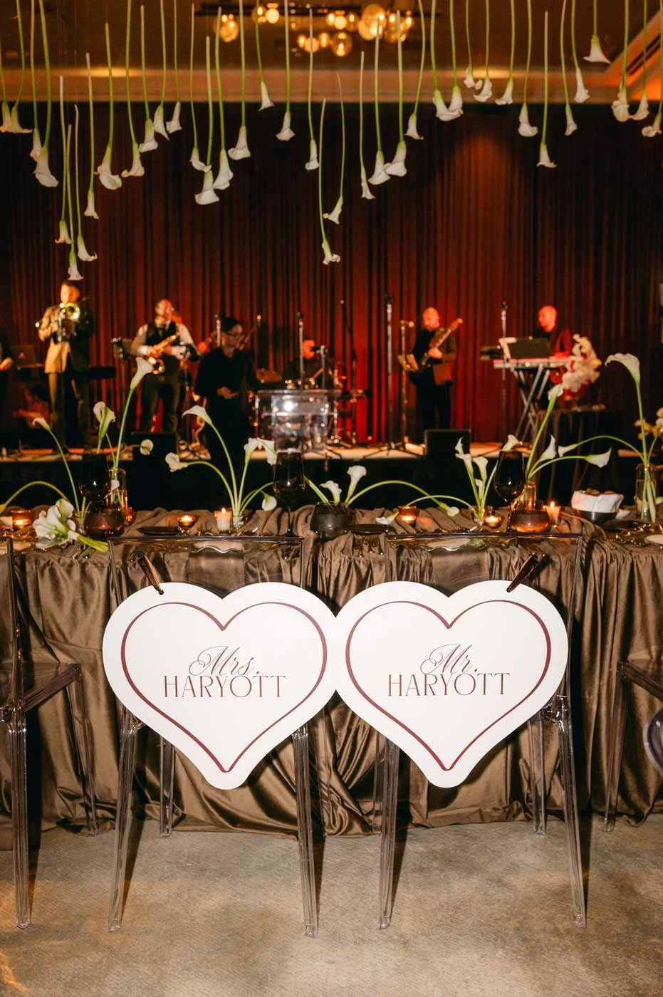 Wedding reception with a stage featuring a live band and decorated tables with floral arrangements, candles, and hearts labeled 'Mrs. Haryott' and 'Mr. Haryott.'