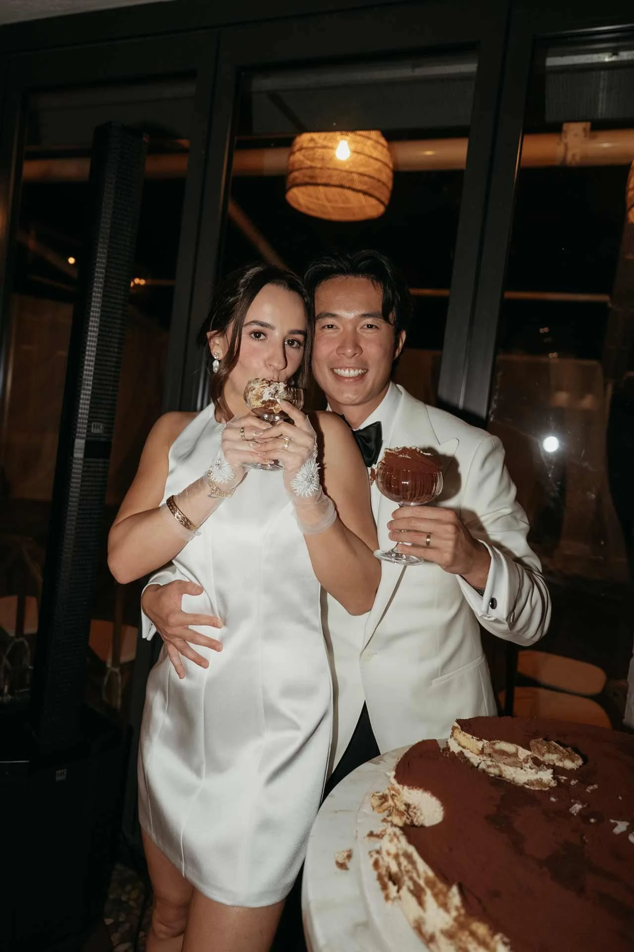 A bride and groom in wedding attire celebrating with dessert and drinks, standing next to a partially eaten chocolate cake.
