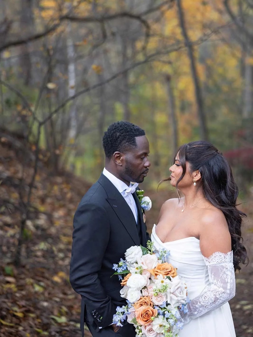 A bride and groom facing each other in a forest setting, dressed in wedding attire, with the bride holding a bouquet of flowers.