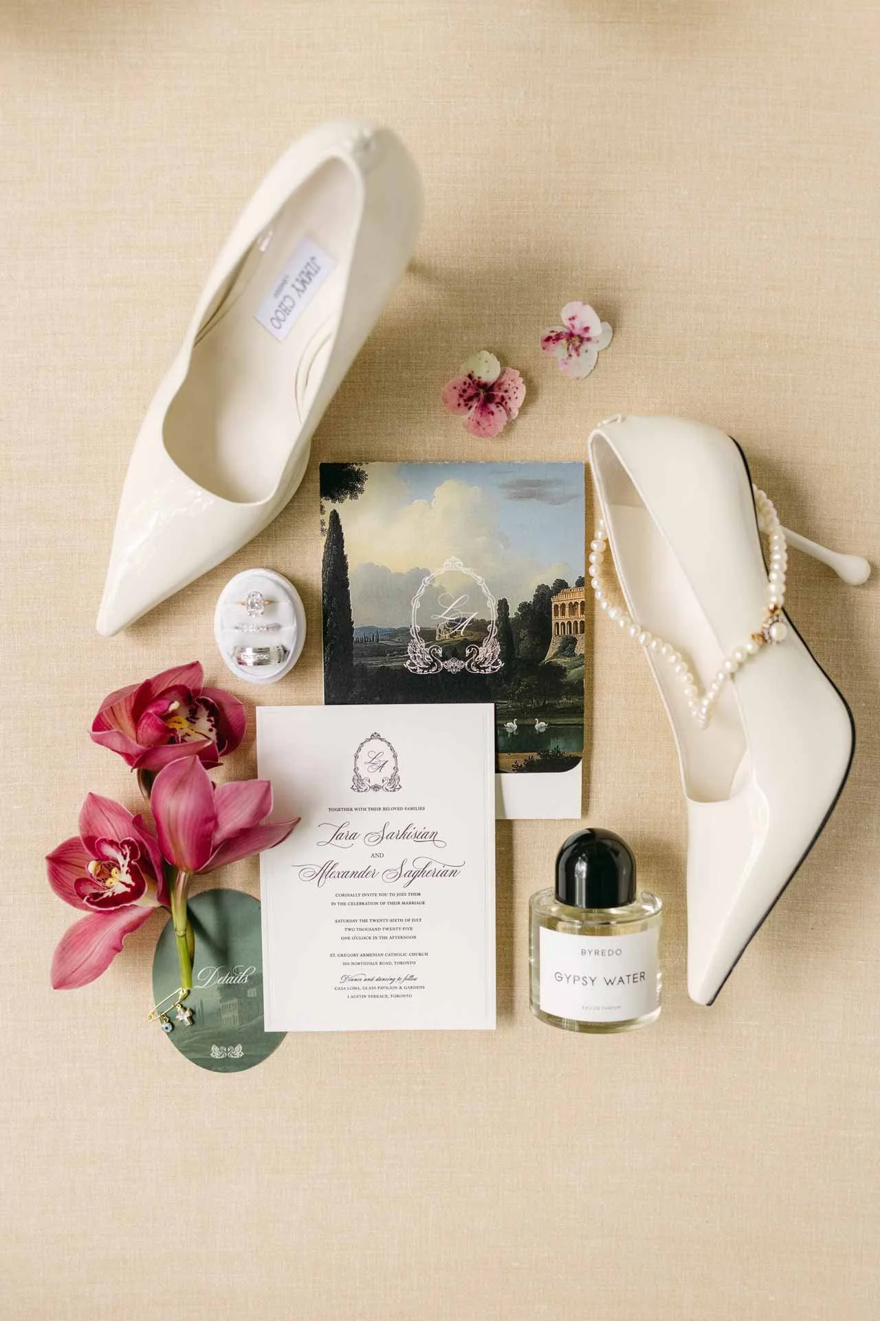 Wedding invitation, white high heel shoes with pearl necklace, pink orchids, a ring, a bottle of Gypsy Water perfume, and a wedding card on a beige background.