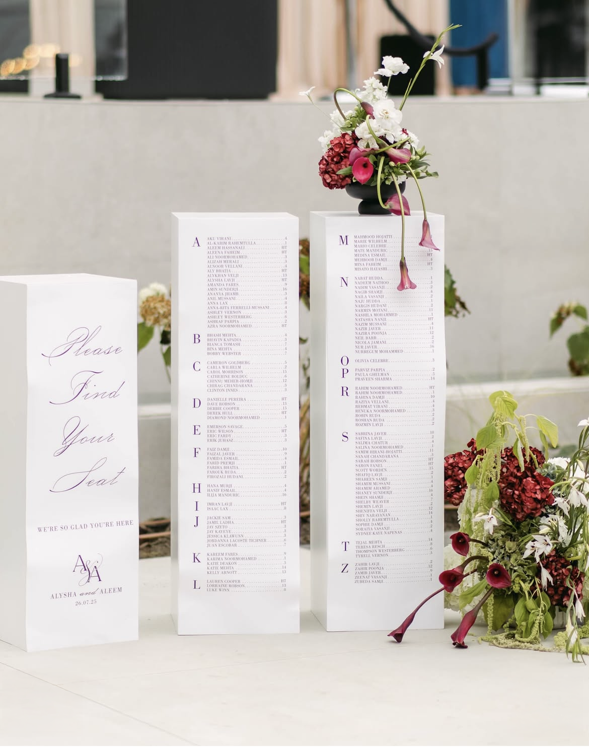 Wedding seating chart display with three vertical panel signs listing guest names alphabetically, decorated with floral arrangements on top and beside.