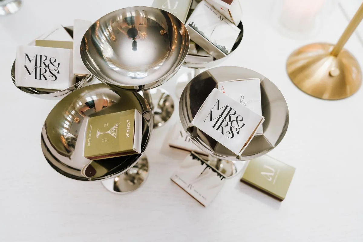 Metal bowls containing wedding and event place cards, with a gold candle holder on a white table.
