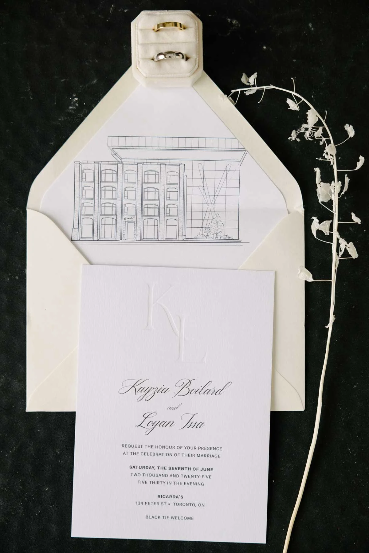 Wedding invitation with details, surrounded by a white envelope, a floral design, and a ring box with two rings, on a black surface.