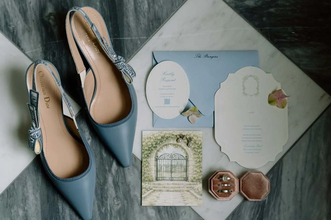 Light blue high-heeled shoes with Dior branding and ribbon straps, wedding invitation cards with floral decorations, a photo of a garden gate, a jewelry box with rings, and an envelope on a marble floor.