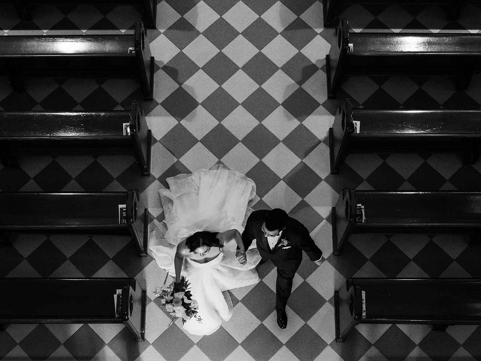 Black and white overhead photo of a bride and groom walking hand in hand down the aisle of a church, surrounded by empty pews.