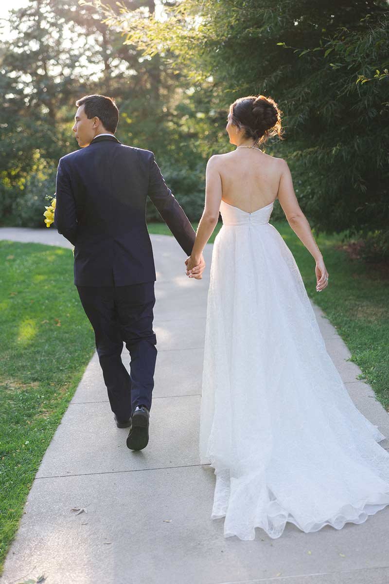 A bride and groom holding hands and walking away on a park sidewalk, surrounded by greenery.