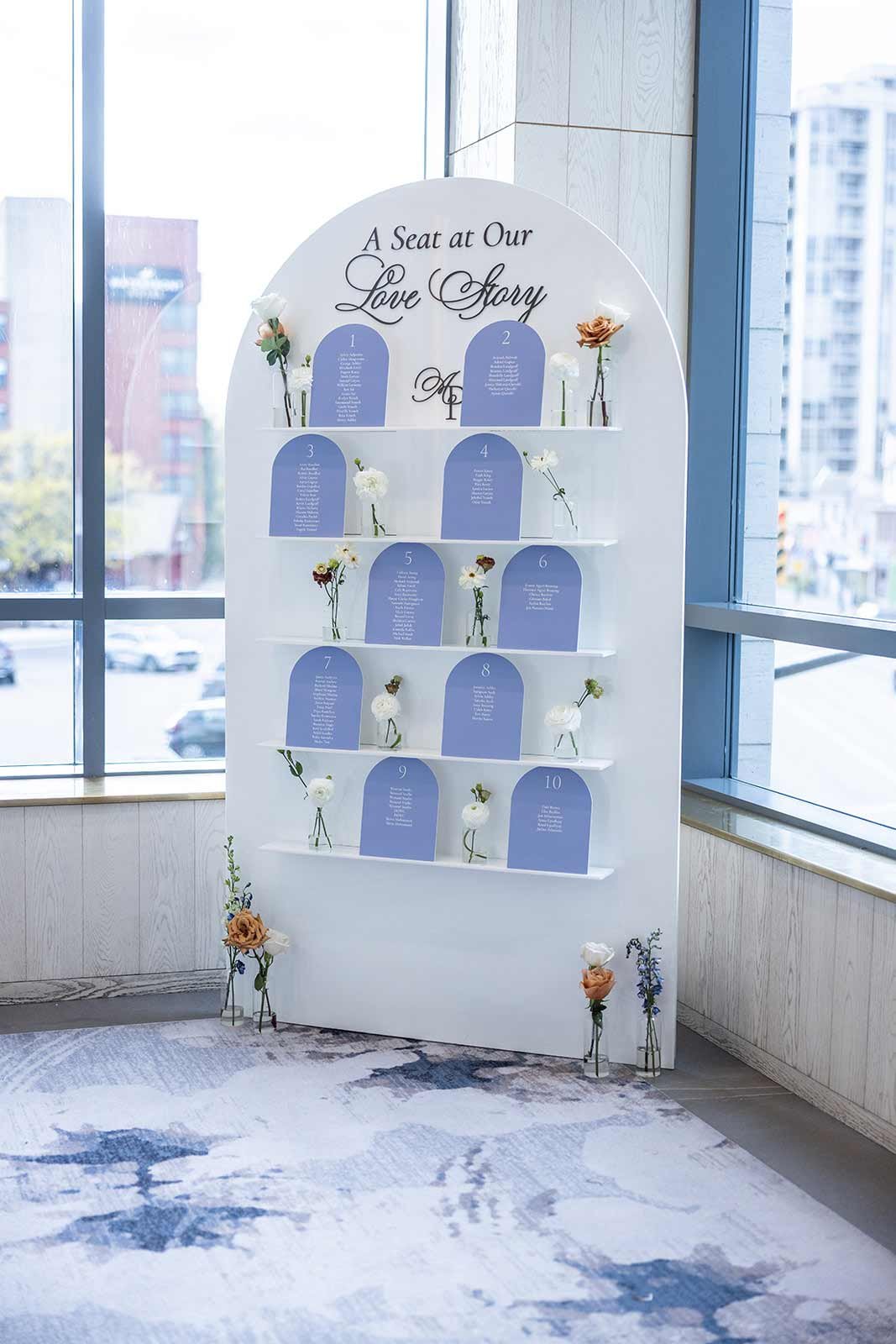 Wedding seating chart display with flowers and table numbers in a hotel or event space, near large windows with city view.