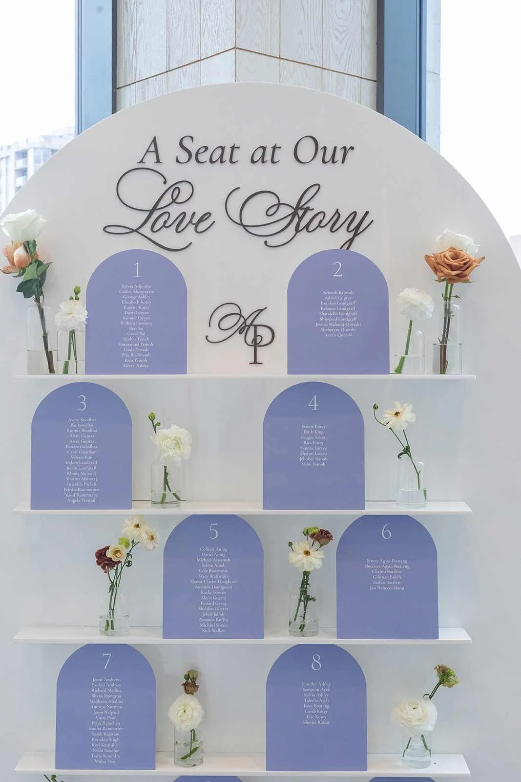 A decorative wedding seating chart with the title 'A Seat at Our Love Story', featuring a list of names organized into eight sections, each with a blue background and white text, surrounded by small glass vases with white, cream, and blush flowers.