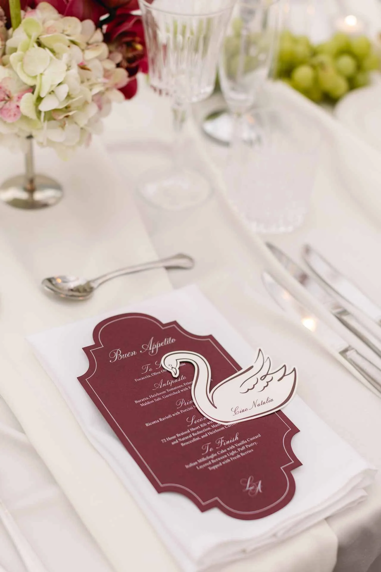 A table setting with a maroon and white menu featuring a swan illustration, surrounded by elegant glassware, silverware, floral arrangements, and a napkin, indicating a formal dining event.
