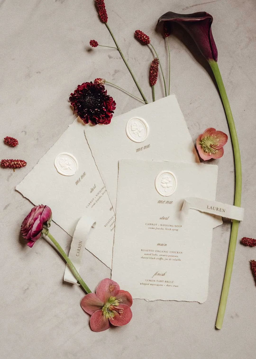 Elegant wedding menu cards surrounded by pink, purple, and red flowers on a light surface.