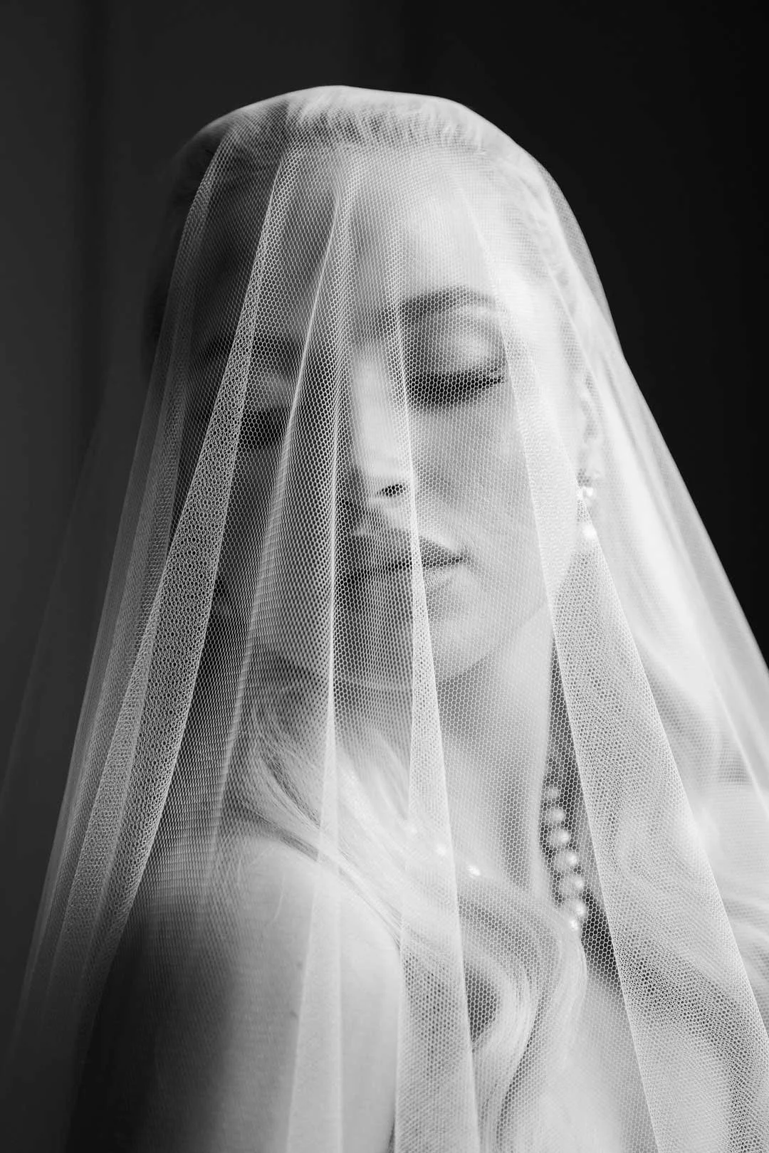 Black and white photo of a woman with a veil over her face, eyes closed, and a necklace.
