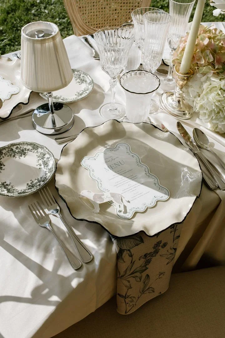 Elegant table setting with fine china, crystal glassware, silverware, a small lamp, and a floral centerpiece, set for a formal meal.