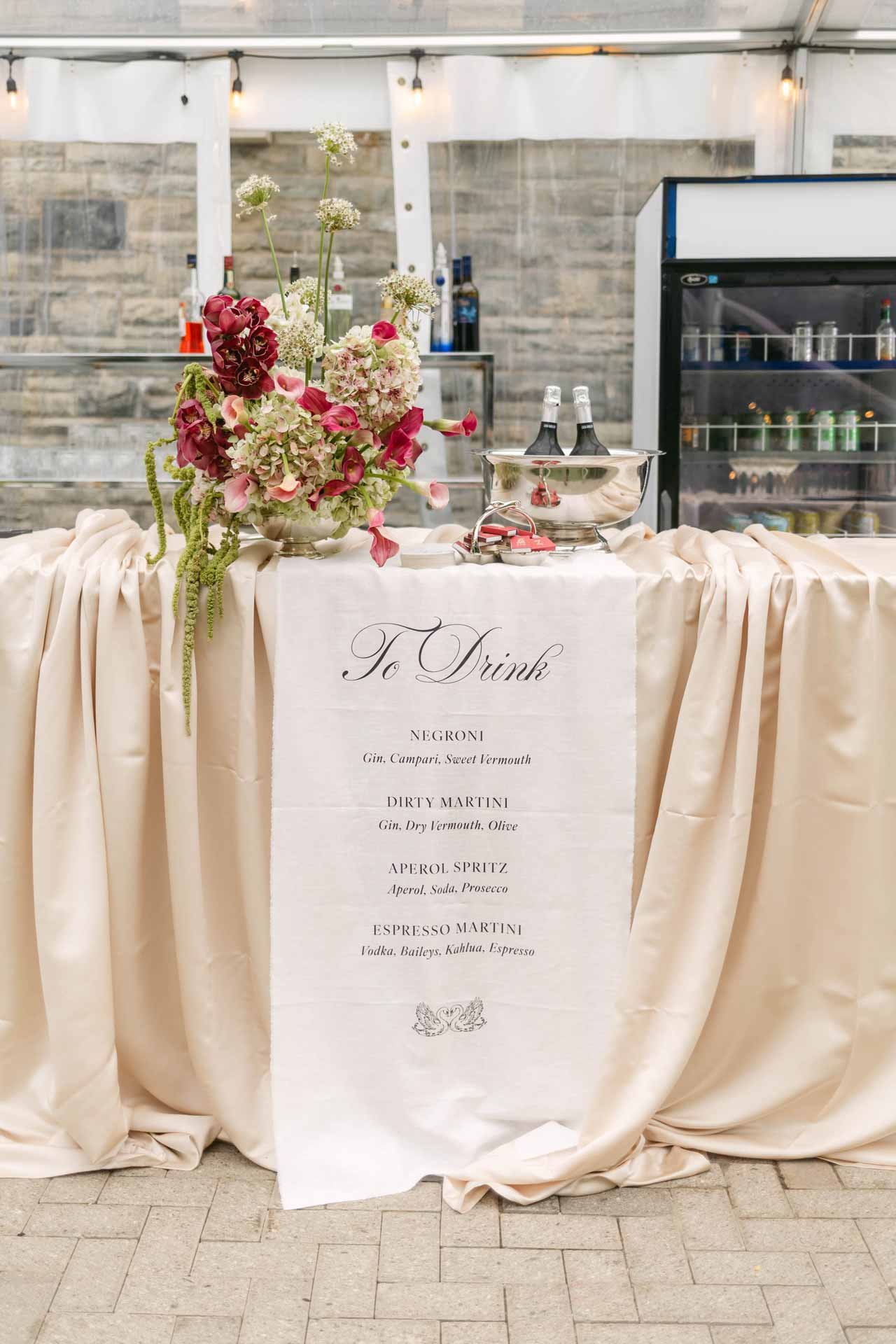 A table with a floral centerpiece, beverage ice bucket, and bar setup in the background, with a menu listing drink options.