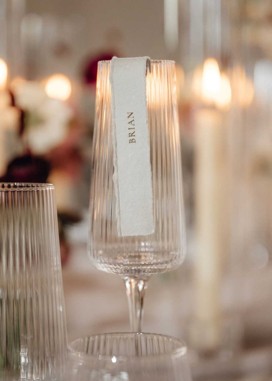 A champagne flute with a paper label that says 'BRIAN' is on a table, with blurred warm lights and decorations in the background.
