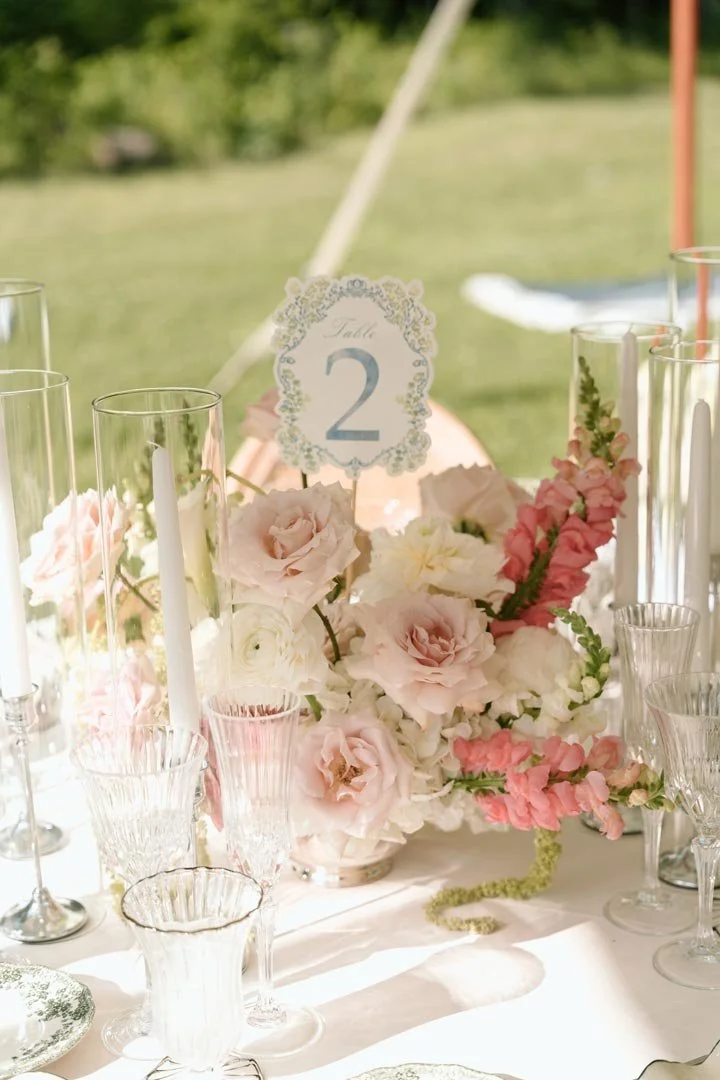 A table centerpiece with pink and white flowers, tall glass candle holders with white candles, and a table number card indicating table 2 at an outdoor event.