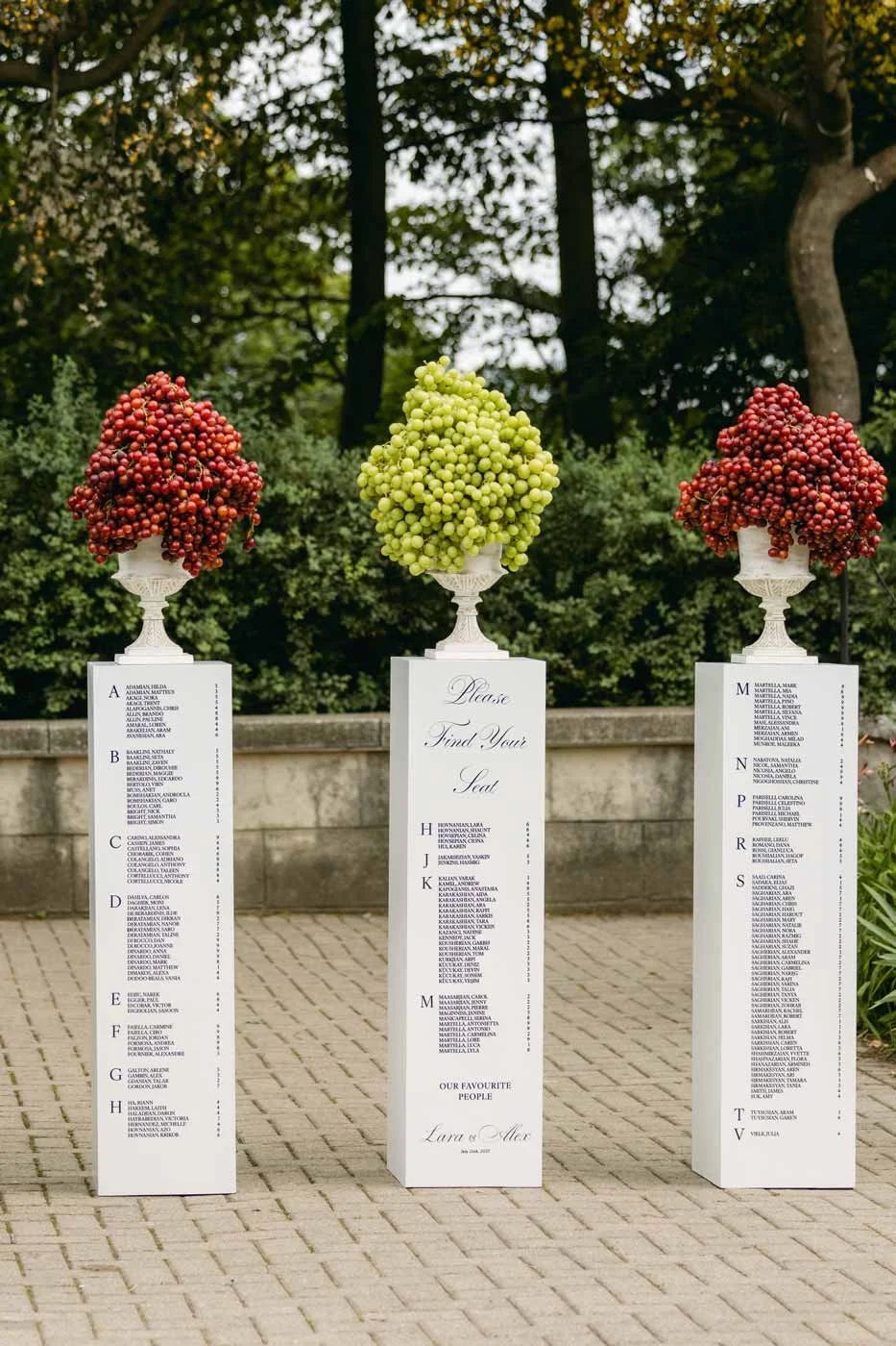 Three white wedding seating chart stands decorated with grape bunches; red grapes on the sides and green grapes in the middle, outdoors with trees in the background.