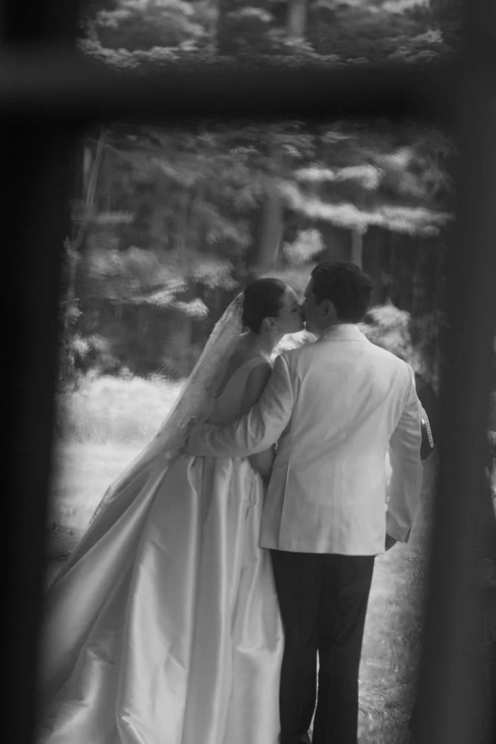 A couple on their wedding day sharing a kiss outdoors, with trees and a lake in the background, seen through a window or frame.