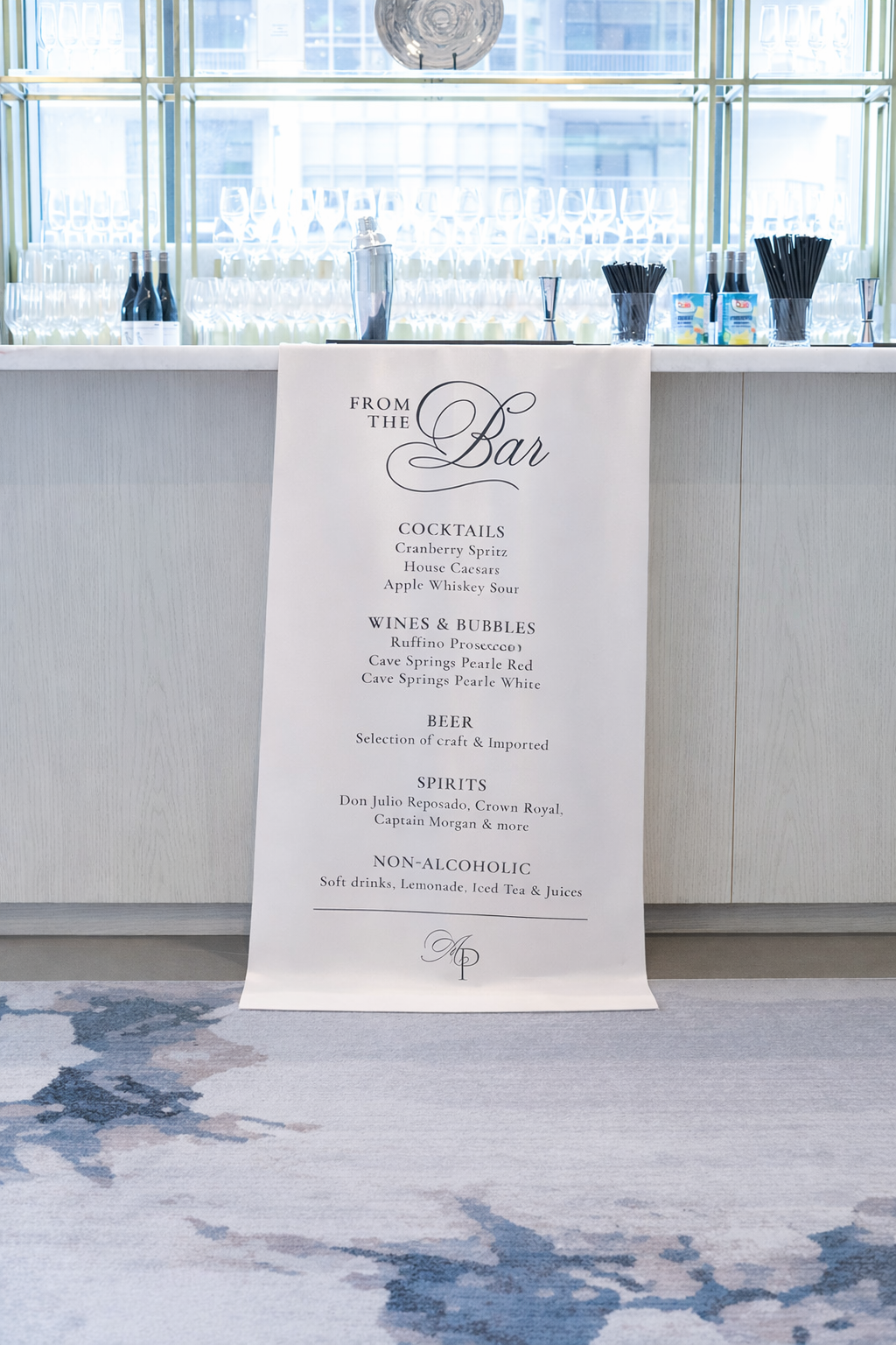 A bar counter with glasses, bottles, and drink straws in front of a large window. A white sign in front lists drink options including cocktails, wines, beer, spirits, and non-alcoholic beverages.