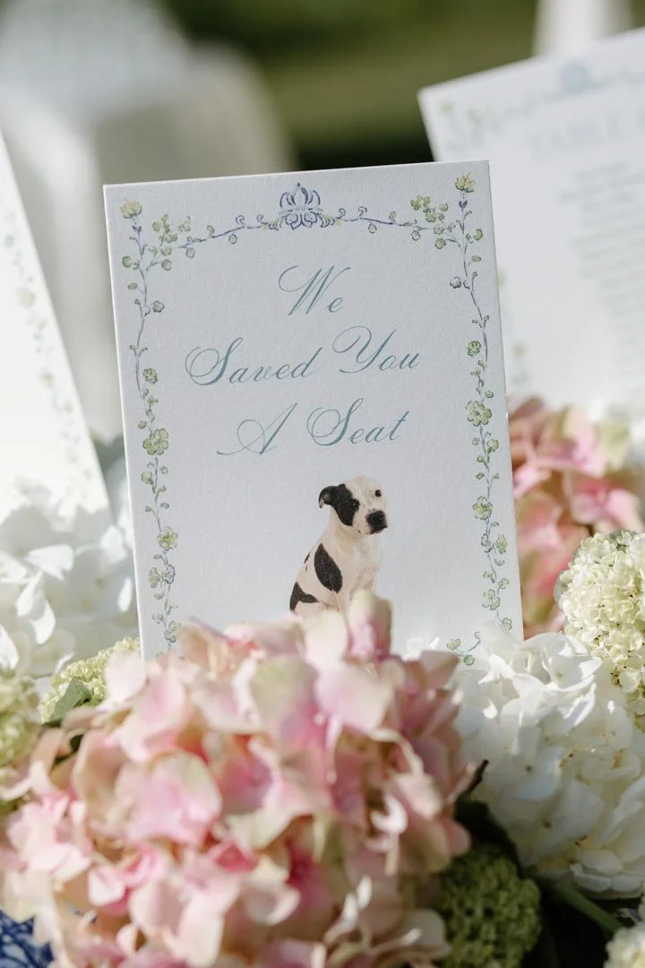A wedding table card with a floral border, a picture of a black and white dog, and the text 'We Saved You A Seat'.