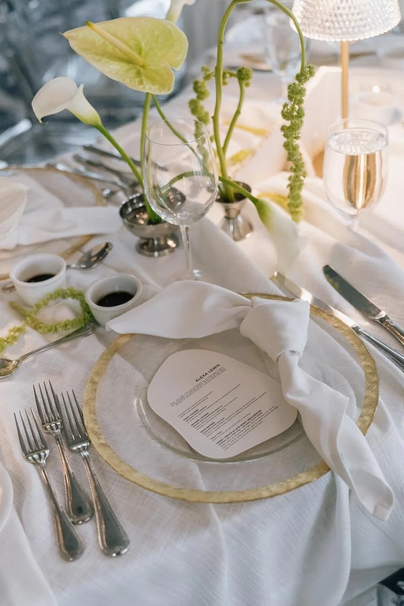 Elegant dining table setting with white tablecloth, gold-rimmed charger plate, folded white napkin, wine glasses, small bowls of sauce, and a floral centerpiece with white calla lilies and green foliage.