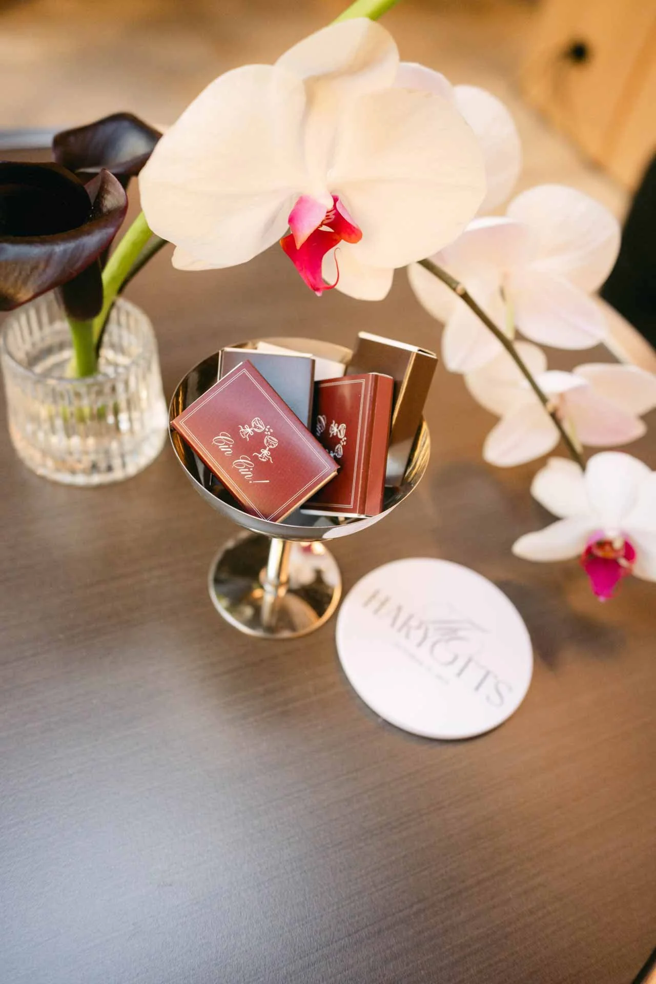 A silver chalice containing small books or cards with French phrases, next to a flower arrangement with white and dark purple orchids on a wooden surface, and a round card with the word 'HAPPINESS' printed on it.