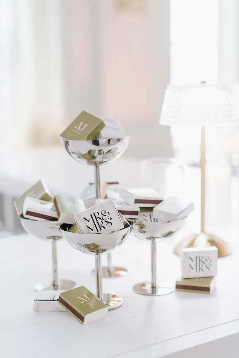 A silver dish stands filled with small match boxes labeled "Mrs & Mrs", with a few boxes scattered around. The setting appears to be indoors, near a window with natural light.