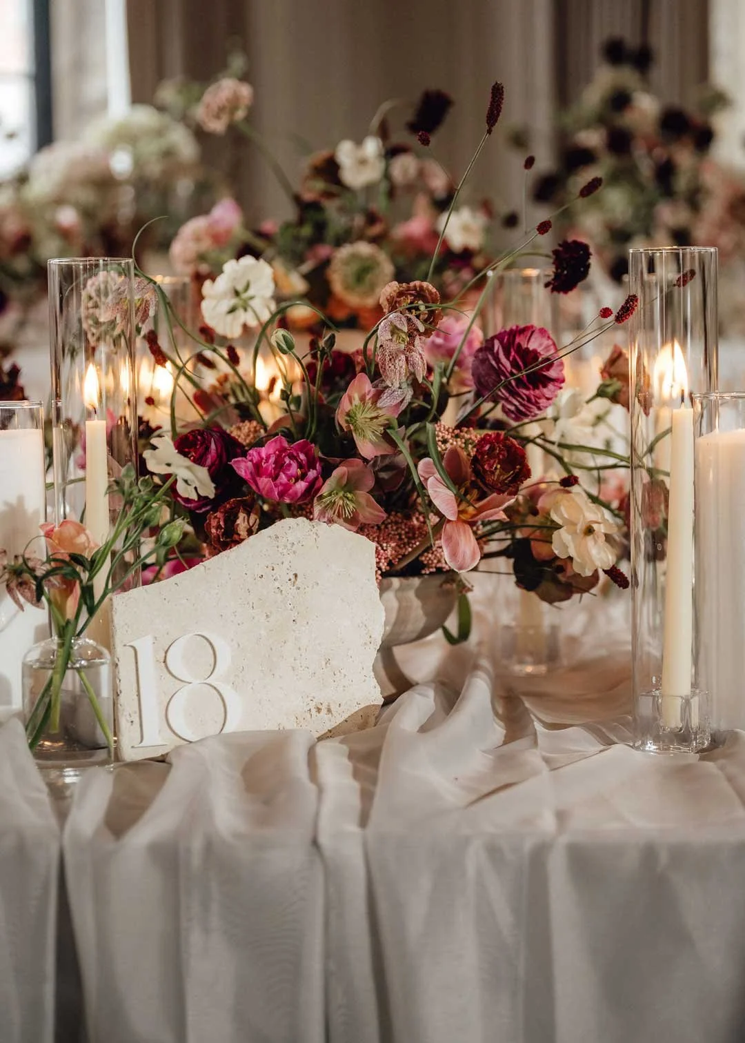 A table centerpiece with a large bouquet of pink, white, and burgundy flowers, surrounded by tall glass candle holders with lit white candles, and a table number 18 card, set on a white tablecloth in a decorated venue.