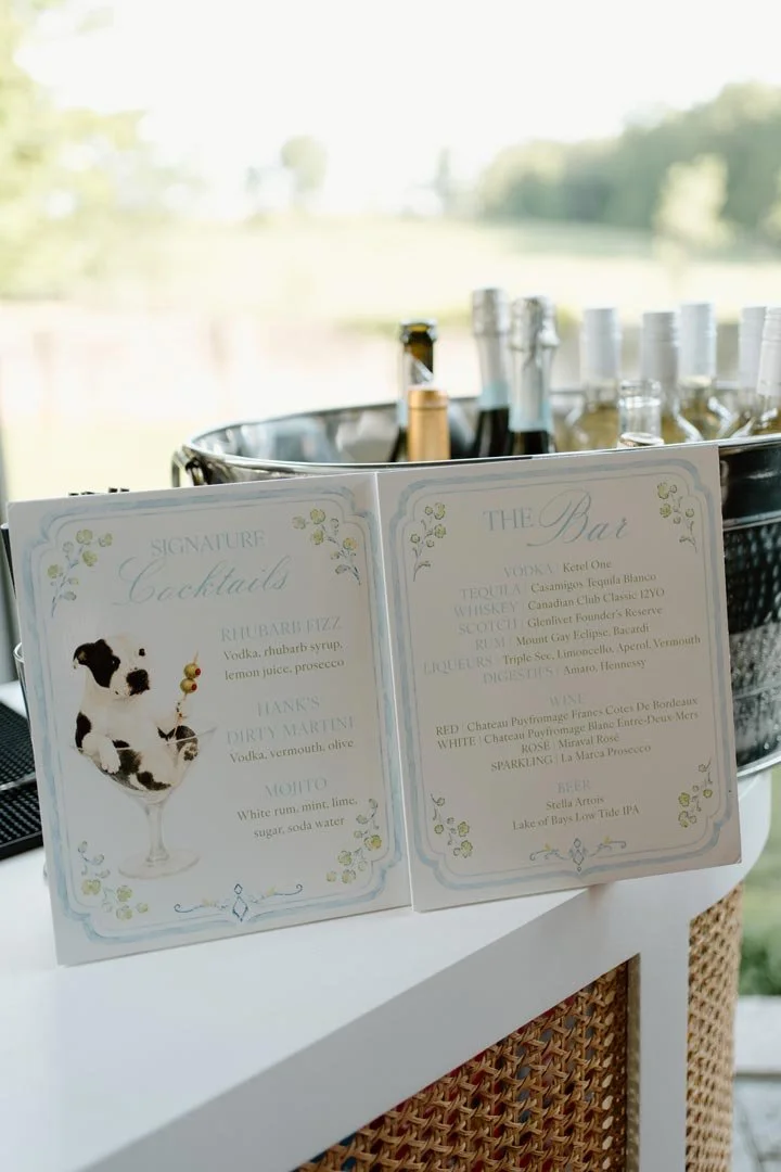 Cocktail menu at a bar with bottles of alcohol in the background, set outdoors with a blurred natural landscape.