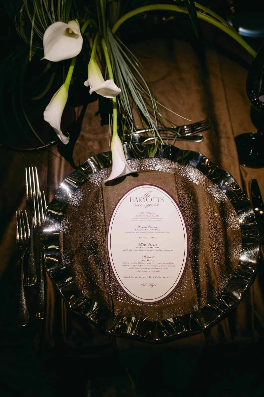 A dinner table setting with a decorative silver charger plate, a printed menu in the center, and a floral arrangement of white calla lilies and greenery.
