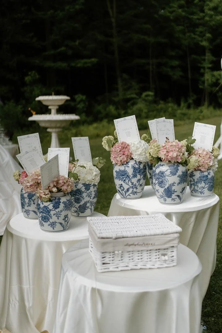 Three small tables draped with white cloths are decorated with blue and white floral patterned vases filled with pink and white hydrangea flowers. Each vase holds a paper card. There is a white wicker basket with napkins or envelopes on one of the ta