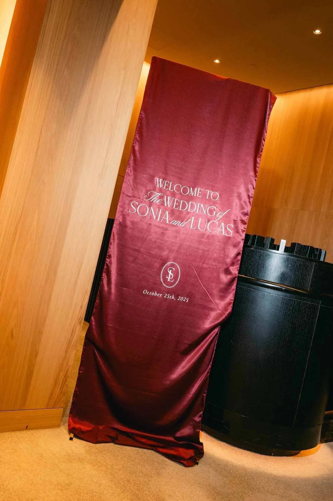A deep red satin wedding sign with white embroidered text reading "Welcome to the wedding of Sonia and Lucas" with the date October 25th, 2025, and a monogram logo in the middle, placed indoors next to a black curved reception desk and wooden wall pa