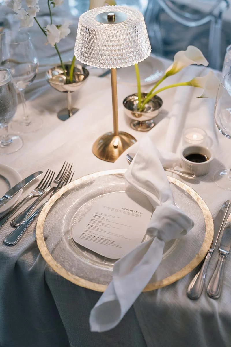Elegant table setting with a silver charger plate, white napkin, silverware, a small candle, a menu, and decorative flowers in cups. A white table lamp provides lighting.