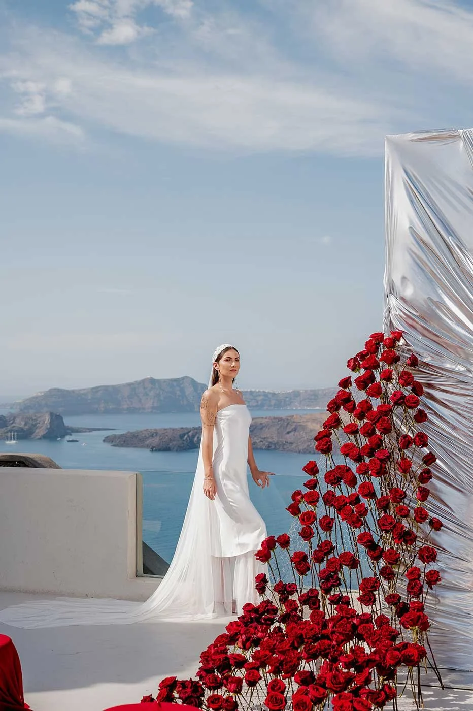 A woman in a white wedding dress and headband stands outdoors on a white platform, with a scenic view of water, cliffs, and islands in the background. A large arrangement of red roses and shiny metallic fabric decorates one side of the scene. Santori