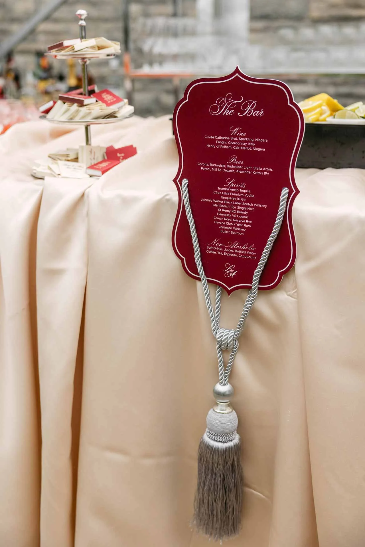 Red decorative sign with white text listing drinks at a bar, hanging from a beige tablecloth with a silver cord and tassel. The sign includes categories such as wine, beer, spirits, and non-alcoholic beverages.