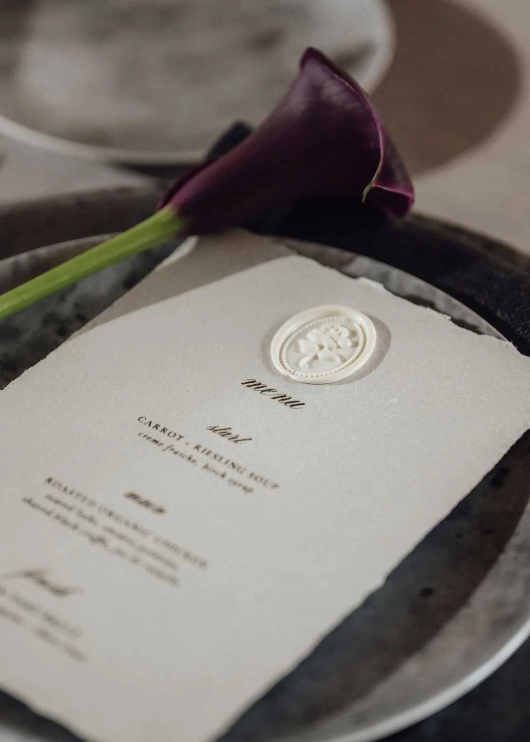 A wedding menu card with a white wax seal and a dark purple calla lily flower on a tray.