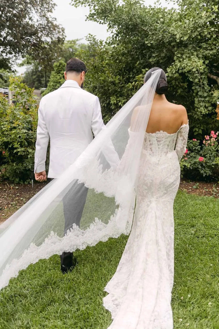 A bride and groom walk together outdoors on a grassy area surrounded by green bushes and trees, with the bride wearing an off-the-shoulder lace wedding dress and veil, and the groom in a white suit.