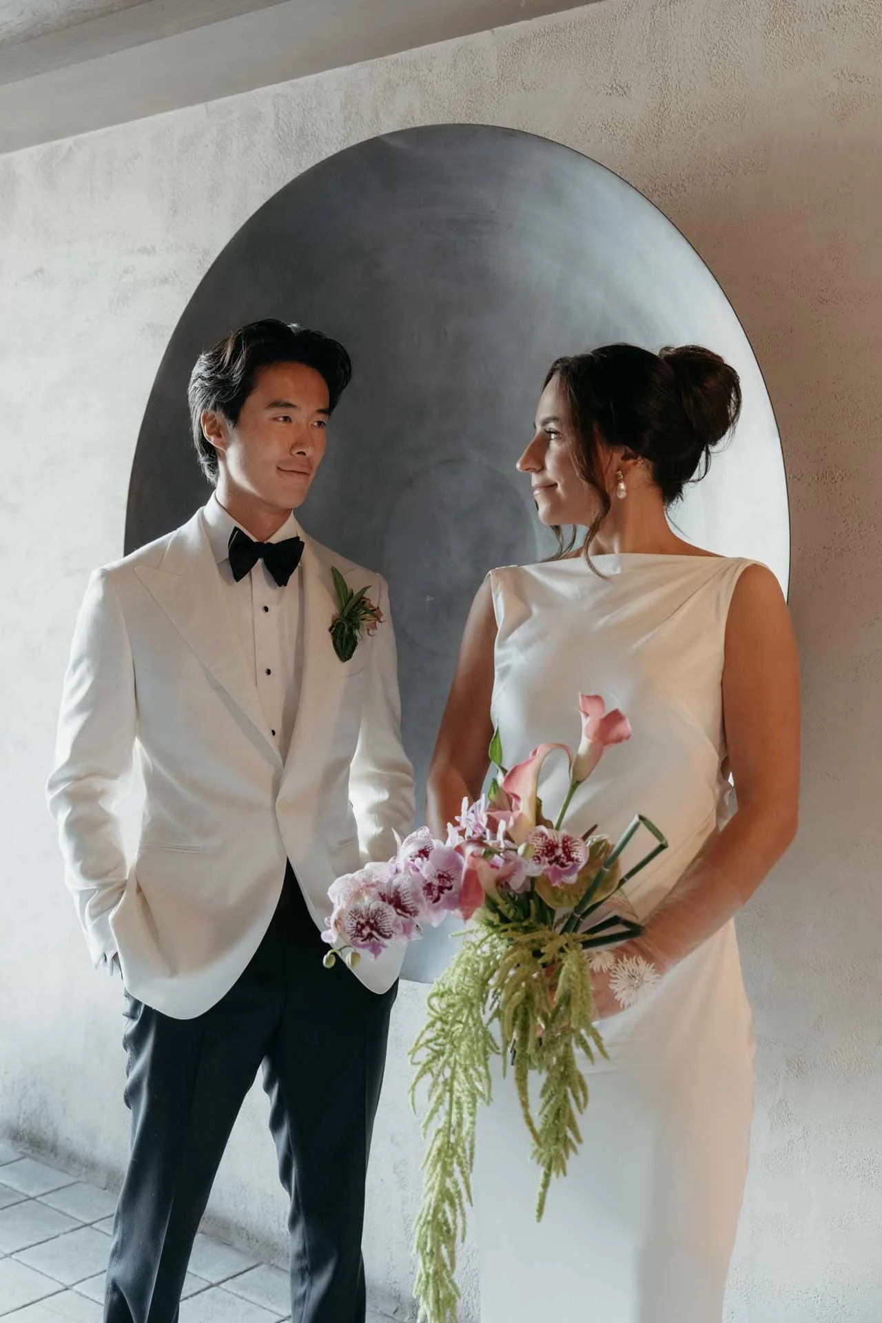 A bride and groom standing indoors, facing each other, with the bride holding a bouquet of pink and white flowers, in front of a round black and gray wall feature.