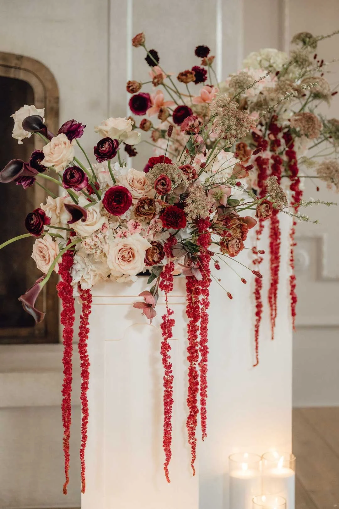 A floral arrangement with pink, red, and dark purple flowers, with hanging red flowers, placed on a white pedestal in an indoor setting.