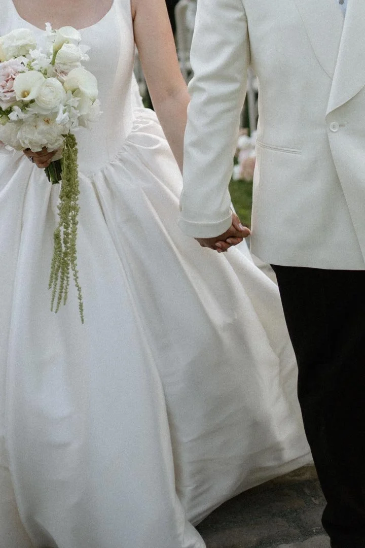 A bride and groom holding hands during a wedding ceremony, focusing on their clothing and hands.