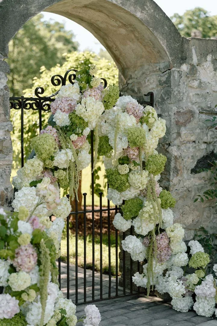 A decorative iron gate adorned with cascading white, pink, and green hydrangea flowers, set against a stone archway with a scenic outdoor background of trees and sky.